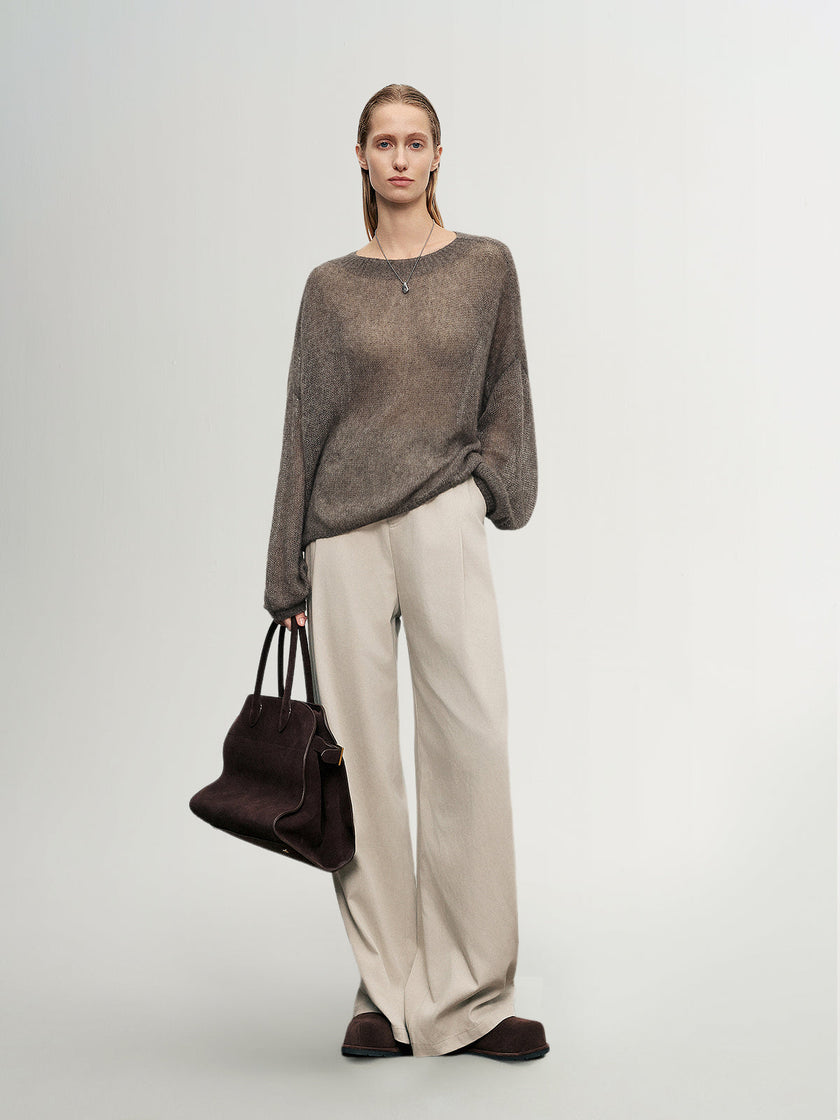 Woman wearing a brown sweater and beige pants holding a brown handbag on a light gray background