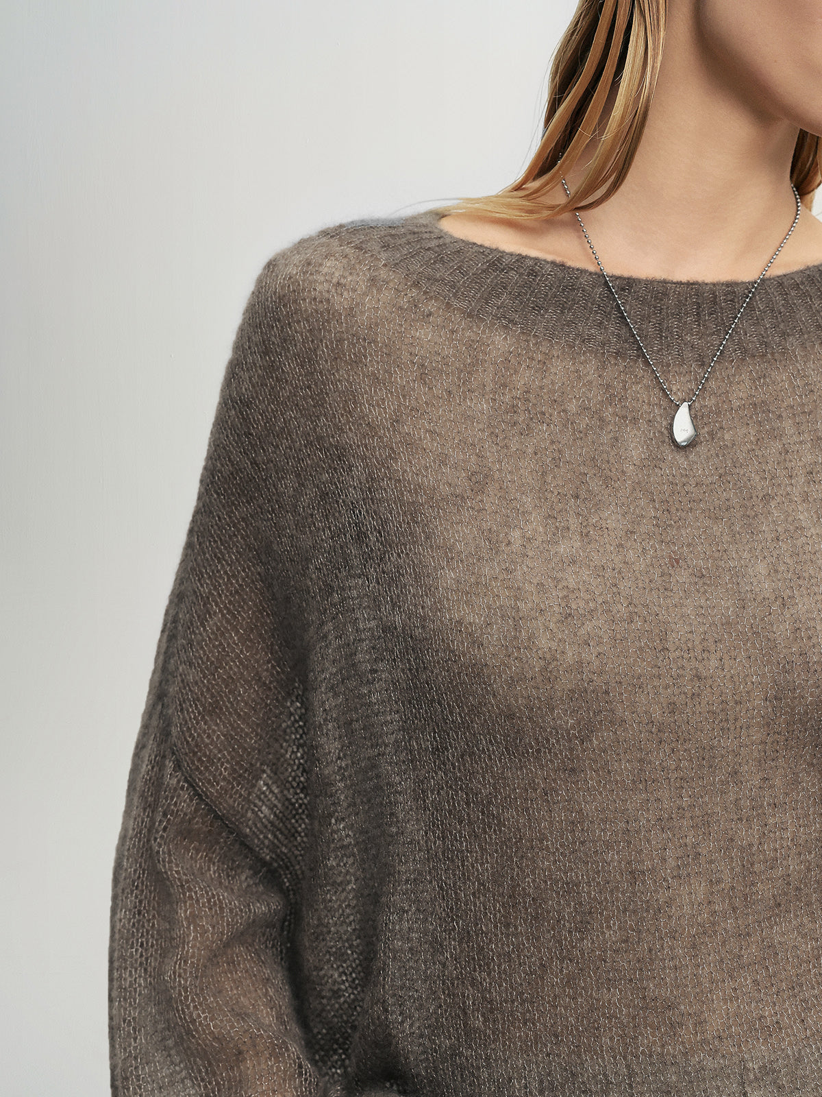 Brown sweater worn by a person with a necklace featuring a teardrop pendant on a plain background