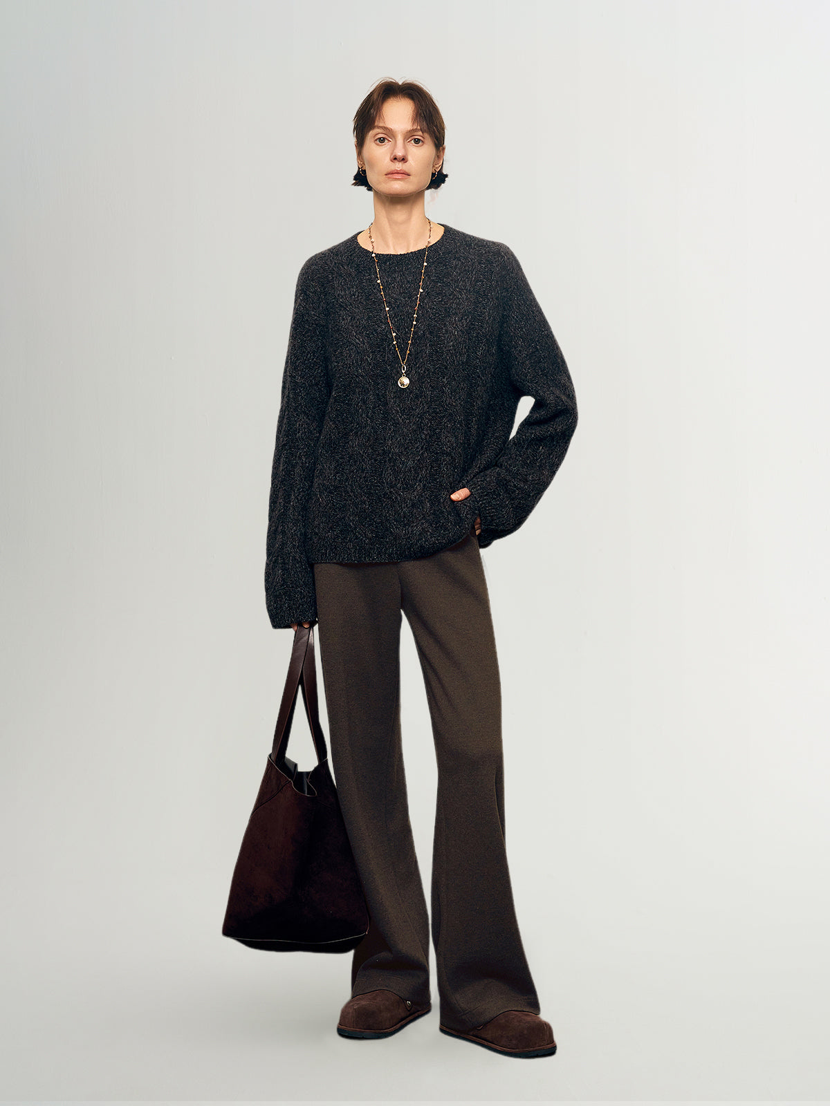 Person wearing a dark sweater and brown pants holding a brown bag on a light gray background