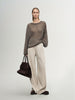 Woman wearing a brown sweater and beige pants holding a brown handbag on a light gray background