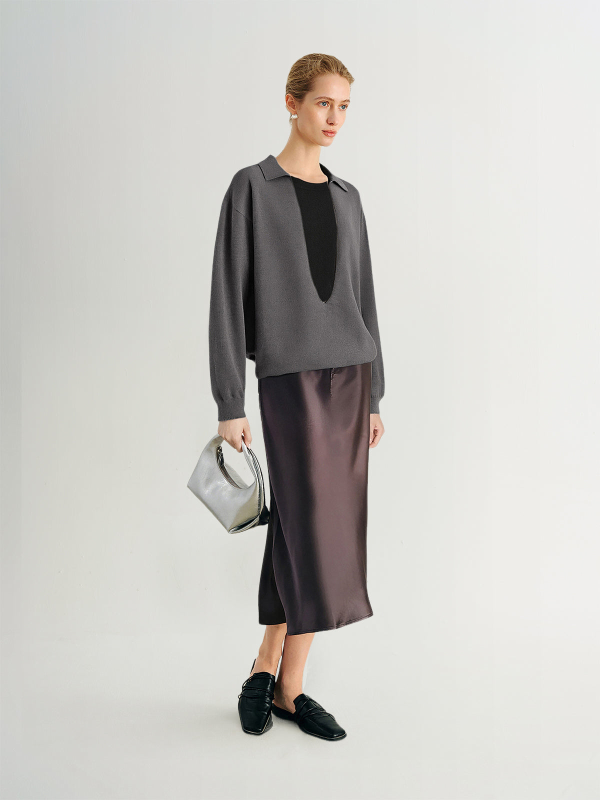 Woman wearing a gray blouse with a black top and satin skirt, holding a gray handbag on a white background.