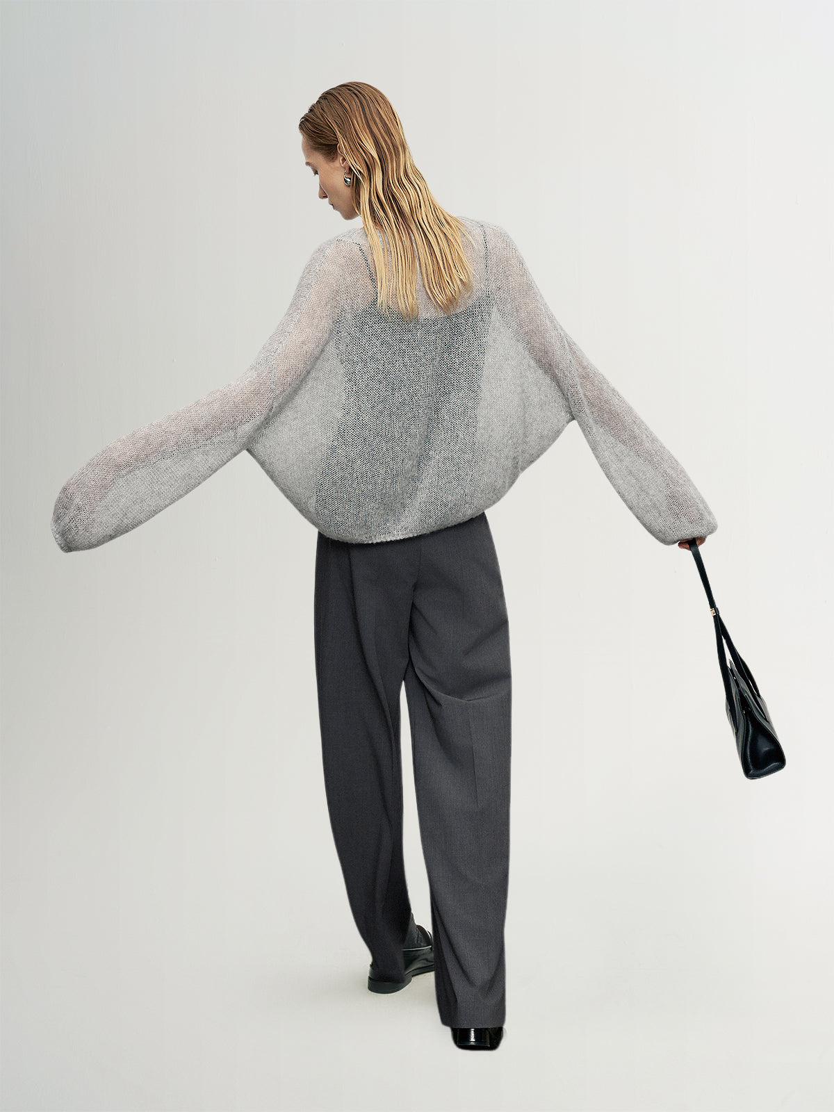Person wearing a gray sweater and black pants holding a black handbag on a white background