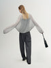 Person wearing a gray sweater and black pants holding a black handbag on a white background